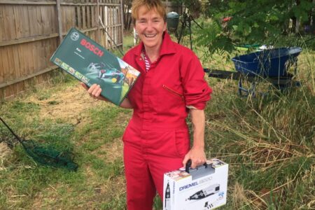 Ruth wears a red boiler suit whilst holding power tools in each hand, standing in the garden