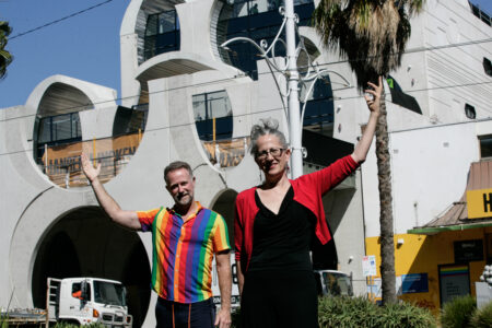 Stuart Kollmorgen from VPC and Karyn Bosomworth from Lord Mayors Charitable foundation stand in front of the pride centre on a sunny day pointing upwards. Stuart is wearing a rainbow shirt and Karyn is wearing black with a red cardigan. Photo Credit: Serge Thomann