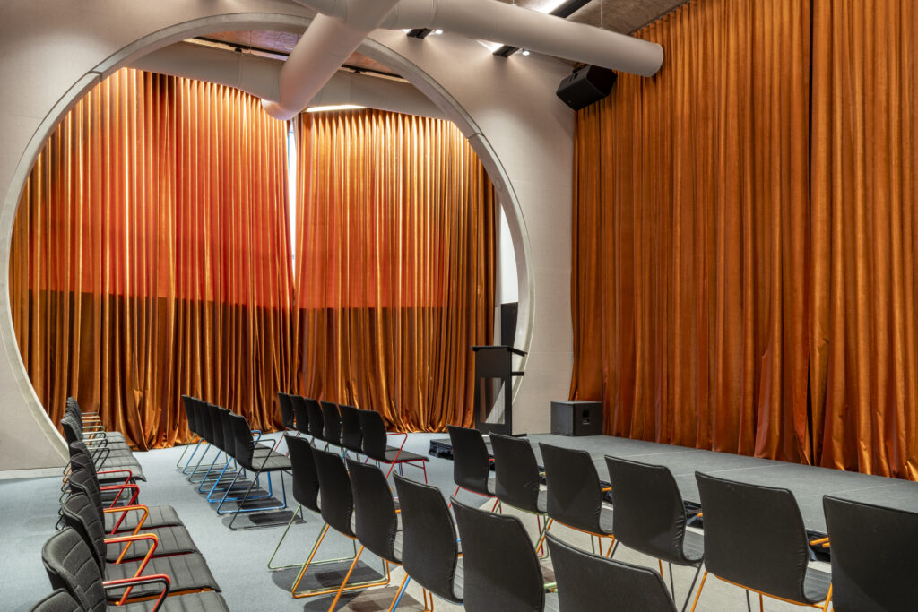 Theatrette with circle framing in front of the large orange velvet curtains blocking a window. There are chairs positioned in rows Infront of a stage as well.