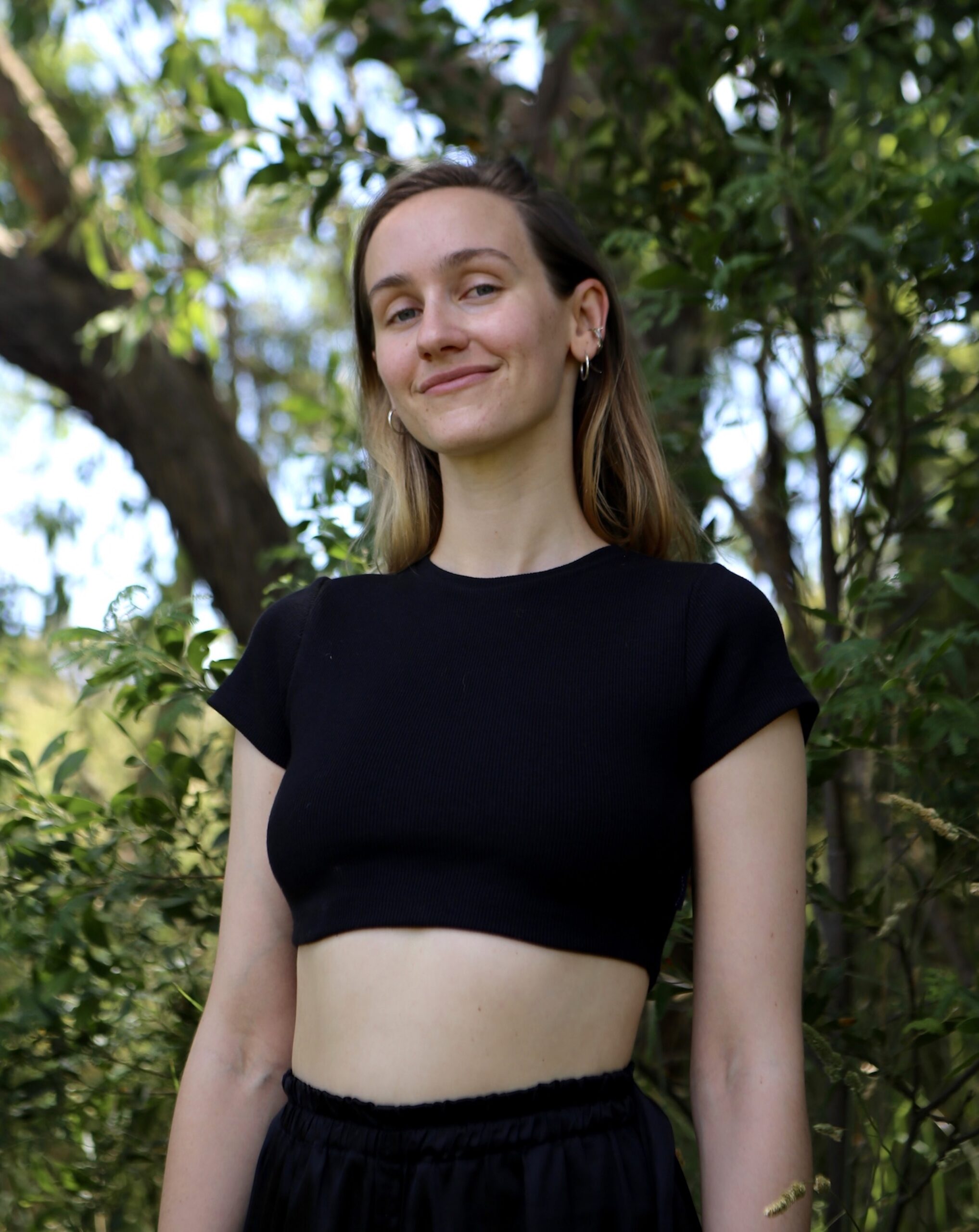 Portrait of Matilda (they/she) surrounded by textured greens of the land with a content expression, wearing a black crop tshirt and high waisted black pants. Portrait of Matilda Brown (they/she)