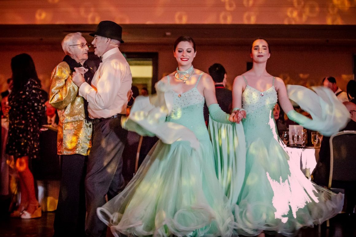 Two queer elders dance next to two young women in ball gowns