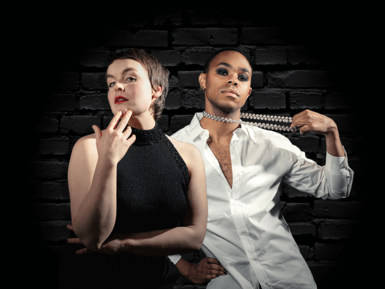 Two people mysteriously pose towards the camera, infront of a black brick wall