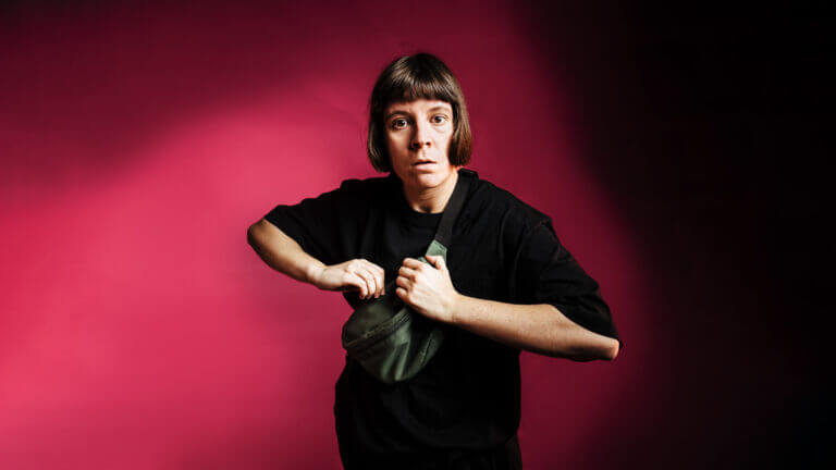 A person with a shocked expression stares at the camera. They stand in front of a pink background.