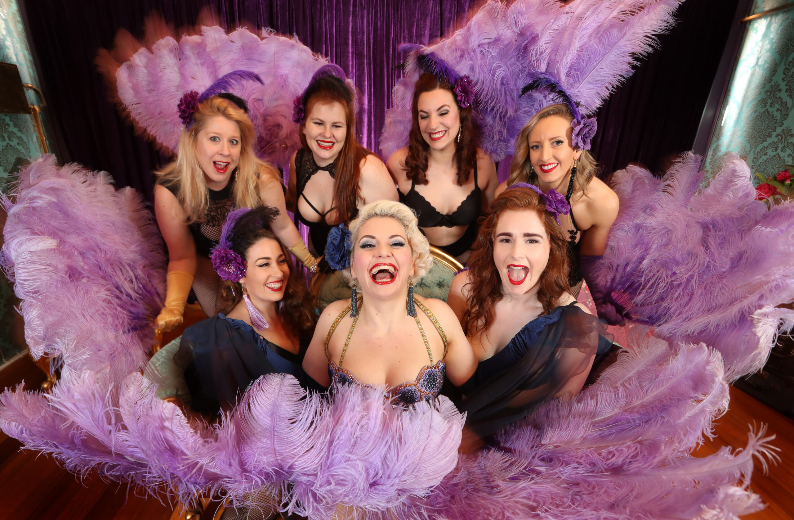 Seven femme-presenting dance3rs encapsulated in purple feather fans.