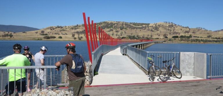 A wide shot of people standing left of a bridge with red accents over a large body of water, extending into a dry landscape