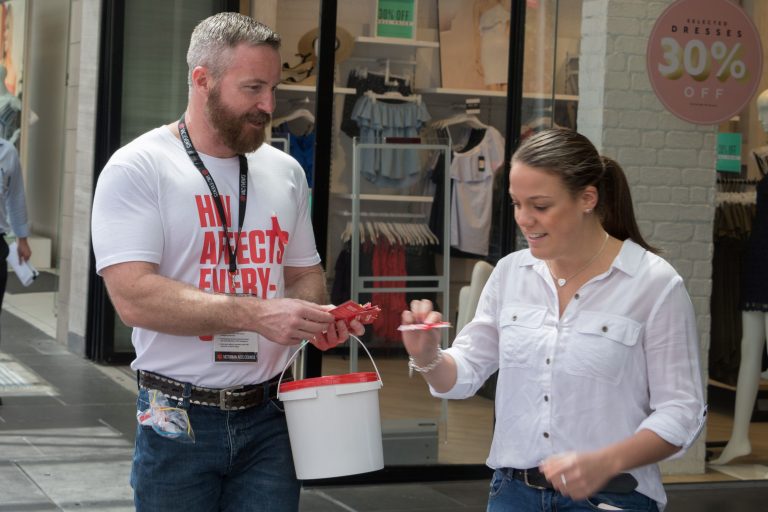 A person in a HIV awareness shirt hands flyers to a pedestrian. They are both smiling.