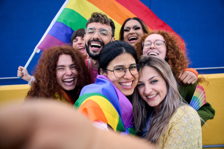 A group of people take a selfie with the pride flag behind them and held by one of the members