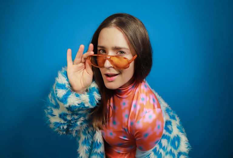 A femme presenting person stares down the barrel of the camera, they hold down their sunglasses, so their eyes are looking just above them.