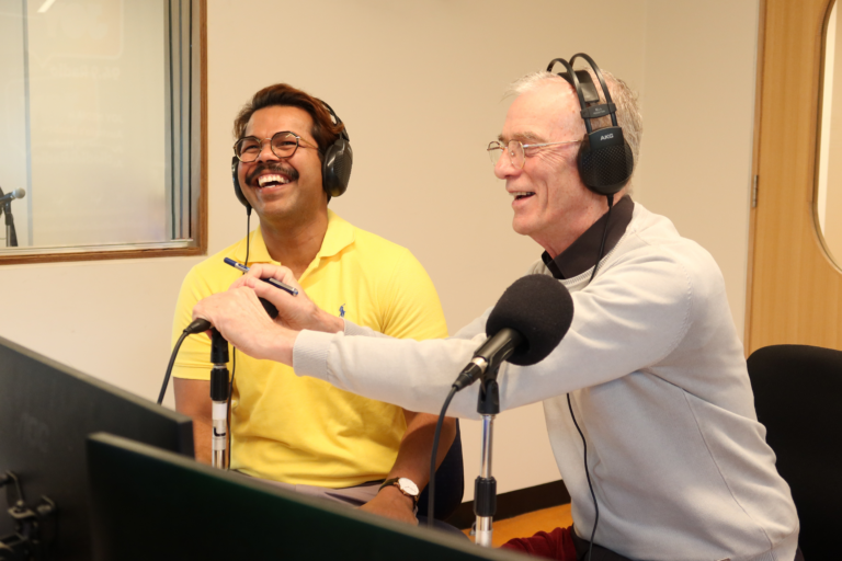 Two people with headphones on smile and speak into microphones