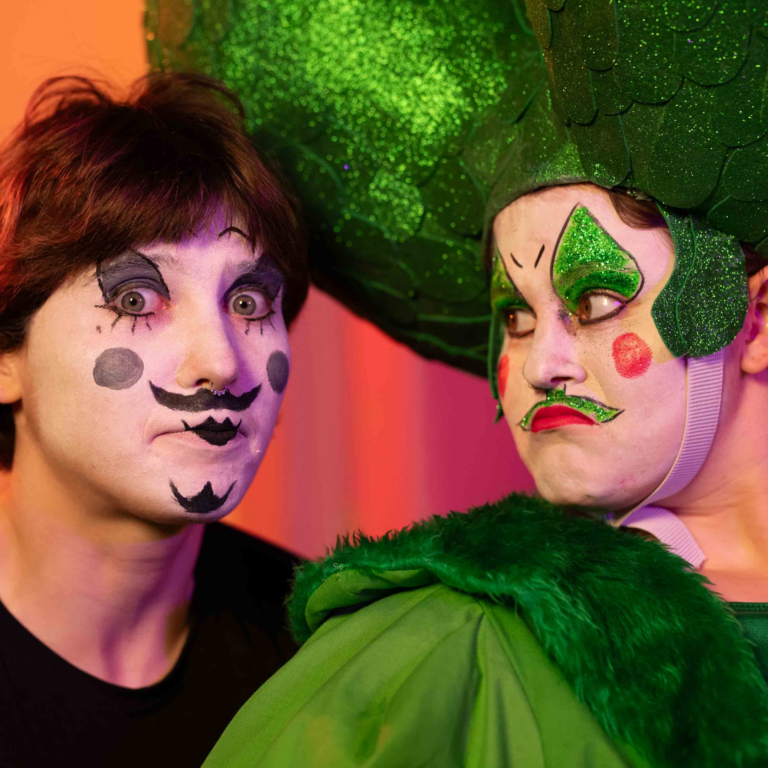 Two people in costumes and clown/drag makeup. They both have large expressions on their face.