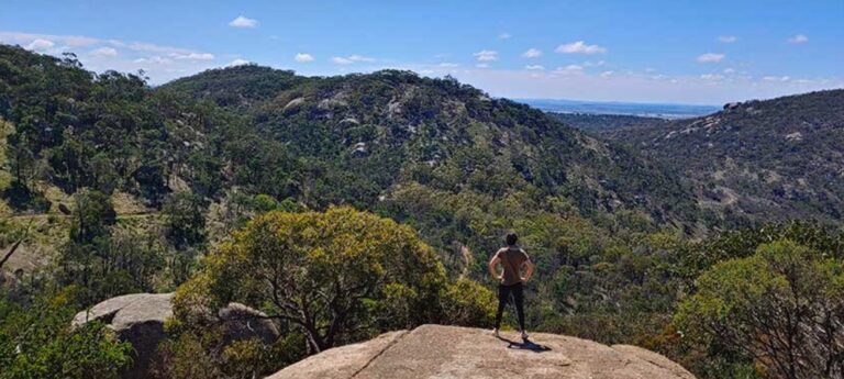 Long shot of the You Yangs - a person stands on a rock looking out towards the trees in the horizon