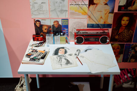 Vintage desk with drawings, tapes, a stereo and posters on a wall.