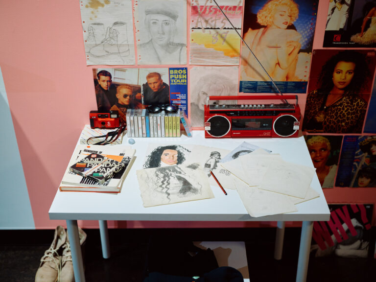 Vintage desk with drawings, tapes, a stereo and posters on a wall.