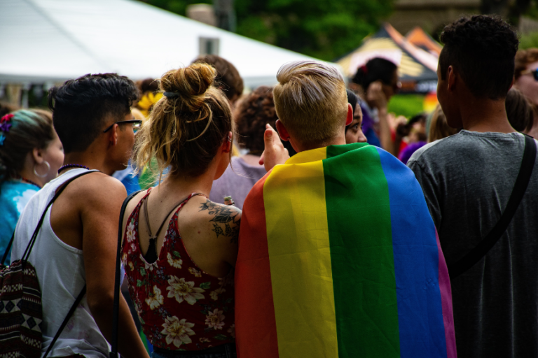 Four people stand with their backs to the camera. One person has wrapped the rainbow flag around their shoulders.