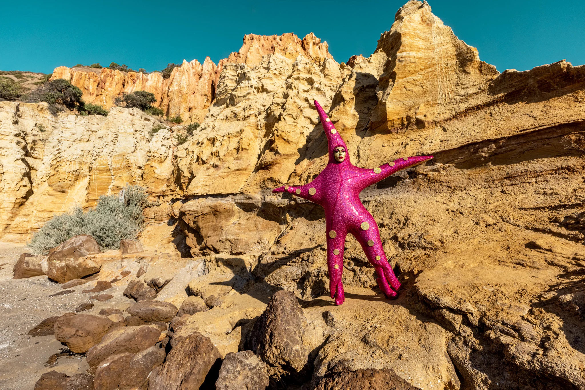 Starfish Sunworship by The Huxleys - a person in an expressionistic costume standing bold on a large rockface