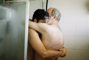 Two masc presenting people hugging in a shower. One person with grey hair has a butterfly tattoo on the back. The image is hazy and the bathroom is full of condensation.