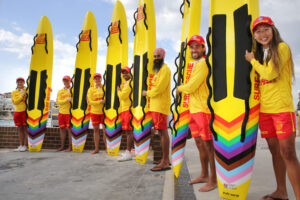 Pride in Sports photo of a group of six people holding surf boards and smiling enthusiastically towards the camera. They all wear Surf Rescue shorts and hold boards with rainbow printing towards the base.