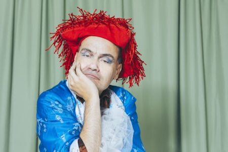 a person stands in a theatre style costume in front of a green backdrop. They rest their head in their hands with one arm crossed around their waist. The person wears a bright red cowboy-style hat.