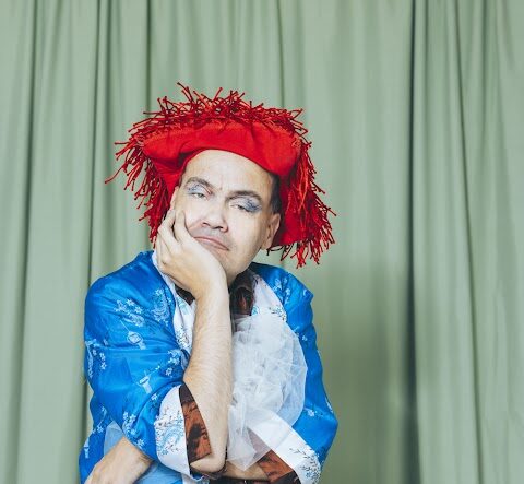 a person stands in a theatre style costume in front of a green backdrop. They rest their head in their hands with one arm crossed around their waist. The person wears a bright red cowboy-style hat.