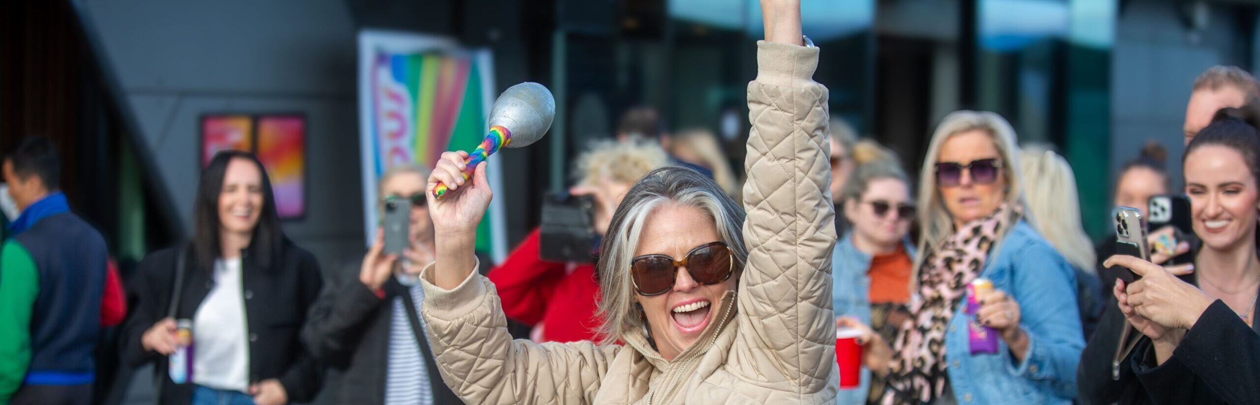 A person with grey, silver hair in a beige jacket holding a pair of maracas amongst a crowd.