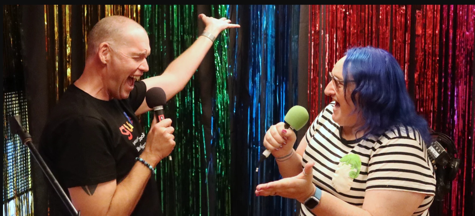 Two people singing in front of a rainbow curtain wall