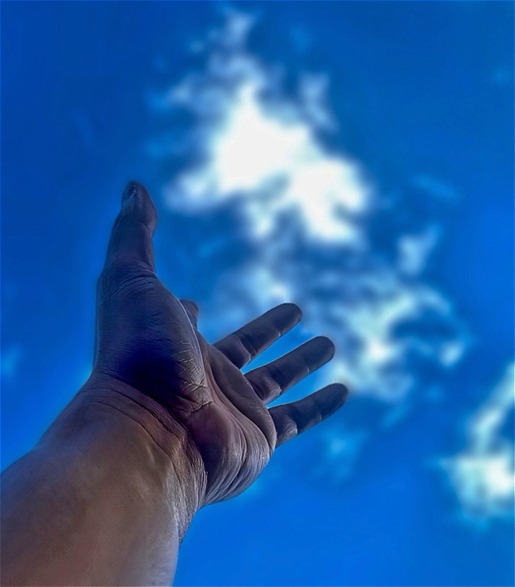 A hand reaches up to the sky with a patch of clouds on blue bright sky