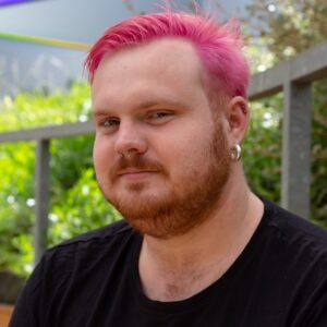 Ryan Hamilton Headshot. He has pink hair, in a crew neck shirt and is looking directly at the camera.